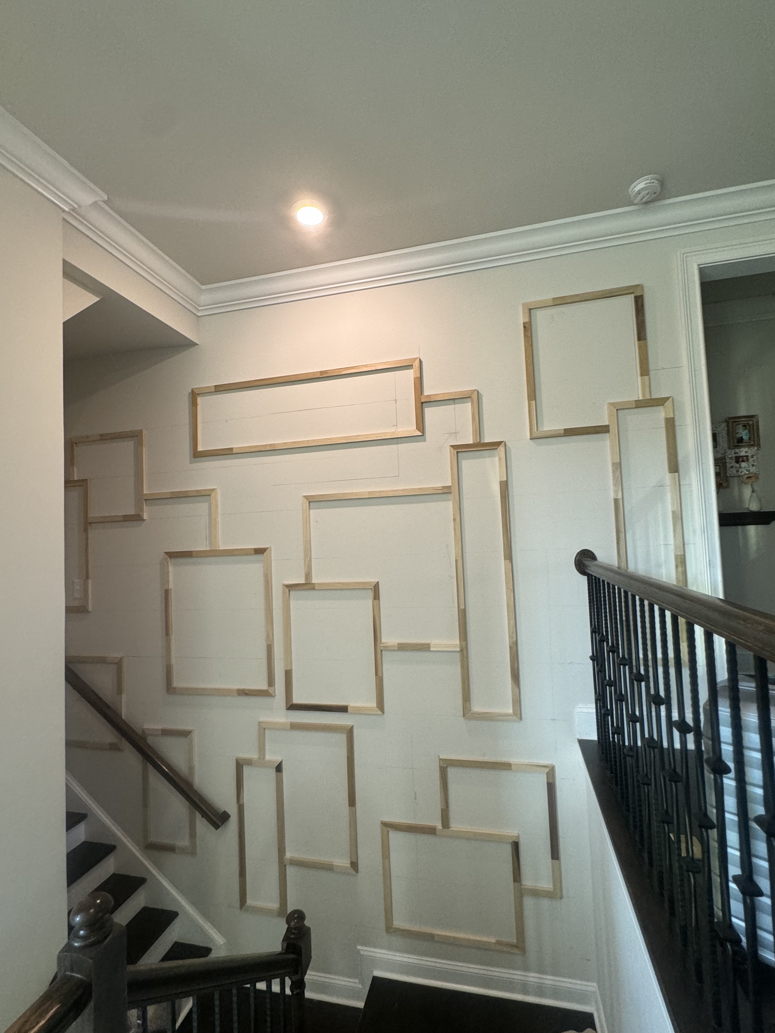 Stairwell with staggered rectangular wood molding on a tall white wall, shown before painting, beside dark stair railings